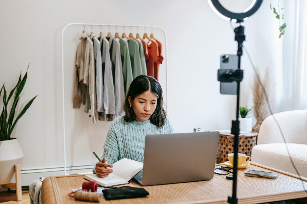 Focused young woman working on laptop in home office, surrounded by clothing rack and camera setup.