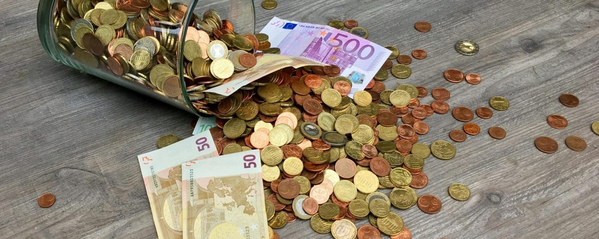 A glass jar with euro coins and banknotes spilled on a wooden surface, representing finance and savings.